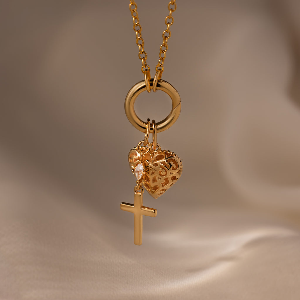 The Diamond Drop Cross Charm in 18K Gold necklace, featuring a heart pendant, is displayed on a soft beige background.