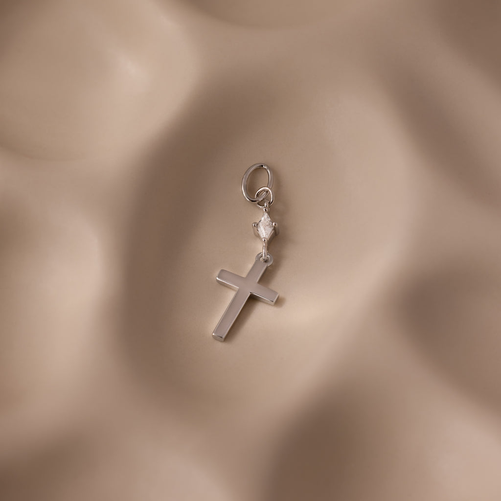 The Diamond Drop Cross Charm features a silver cross with a small heart detail and a subtle diamond accent, set against a soft, wavy beige background.