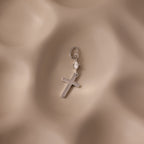 The Diamond Drop Cross Charm features a silver cross with a small heart detail and a subtle diamond accent, set against a soft, wavy beige background.