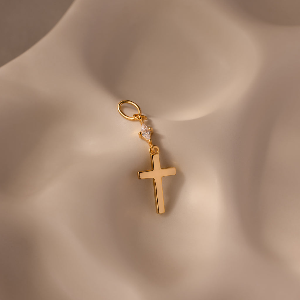 The Diamond Drop Cross Charm in 18K Gold features a delicate diamond accent and is elegantly displayed on a softly textured beige surface.