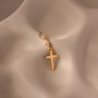 The Diamond Drop Cross Charm in 18K Gold features a delicate diamond accent and is elegantly displayed on a softly textured beige surface.