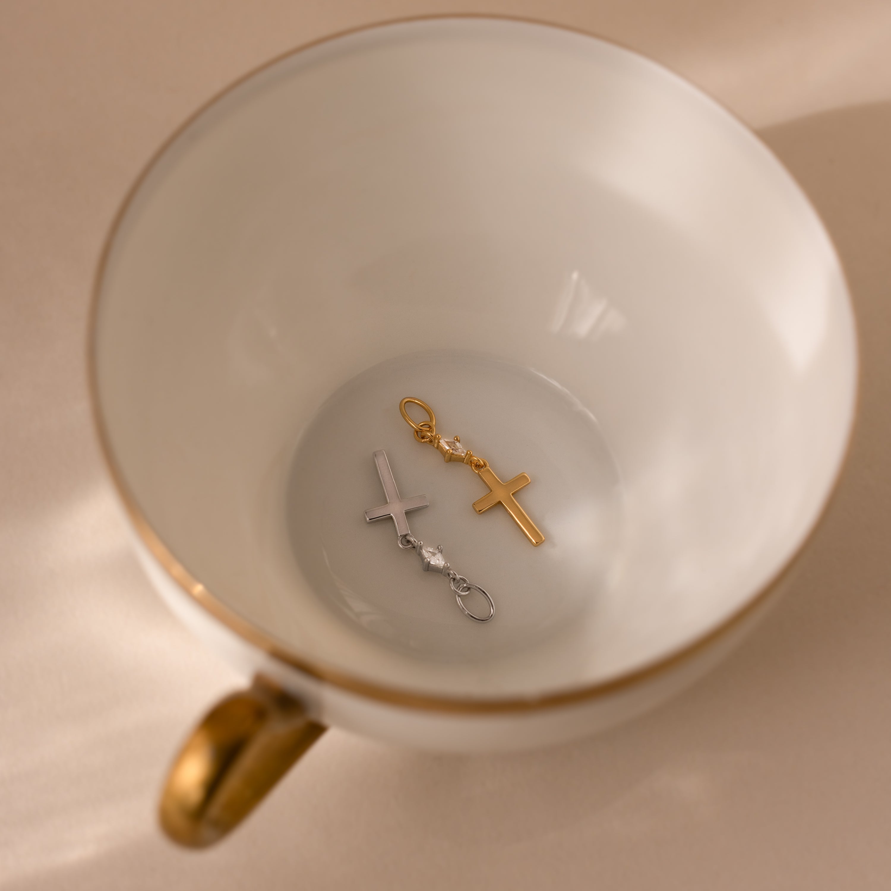 The Diamond Drop Cross Charm, available in both gold and silver with a delicate diamond accent, is displayed inside a white teacup featuring a gold rim and handle.