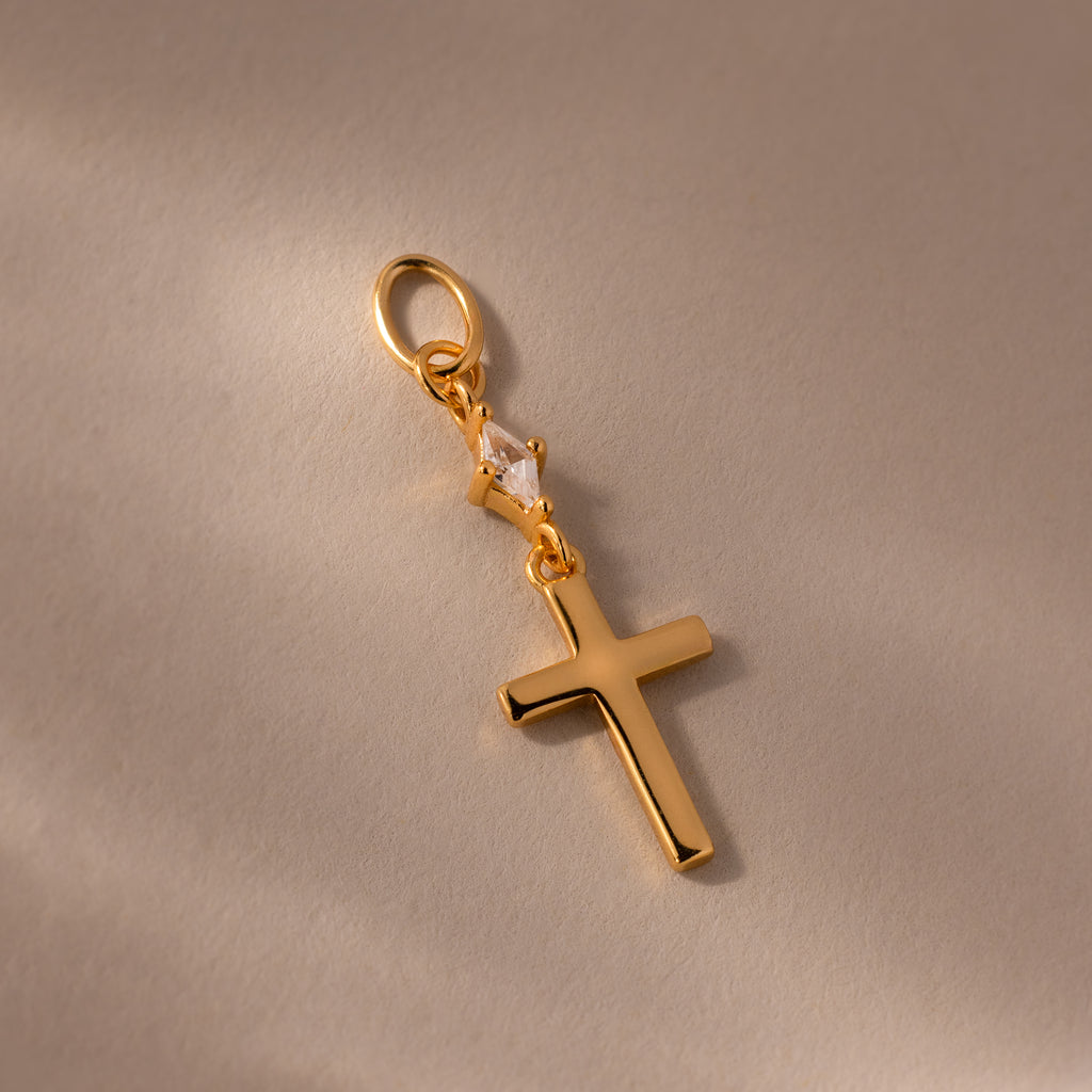 Diamond Drop Cross Charm in 18K Gold, featuring a small diamond accent, displayed on a light beige background.