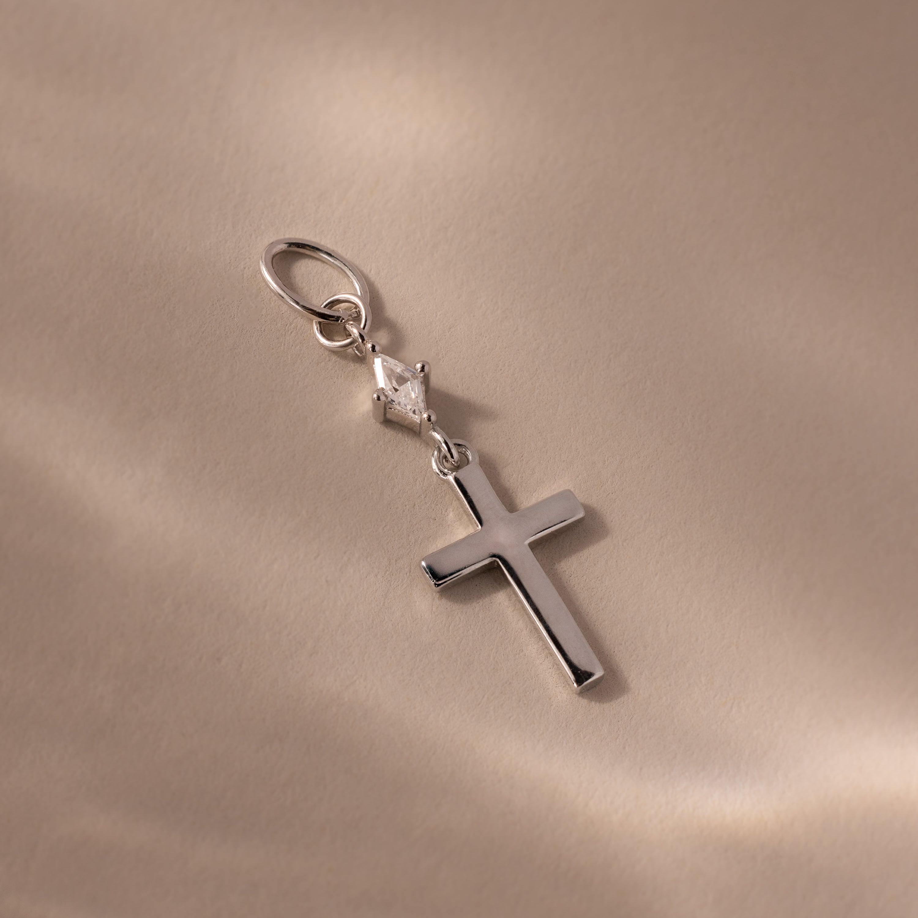 Delicate Diamond Drop Cross Charm in Silver featuring a small, sparkling diamond detail, shown on a beige surface in soft light—a stunning accessory for any occasion.