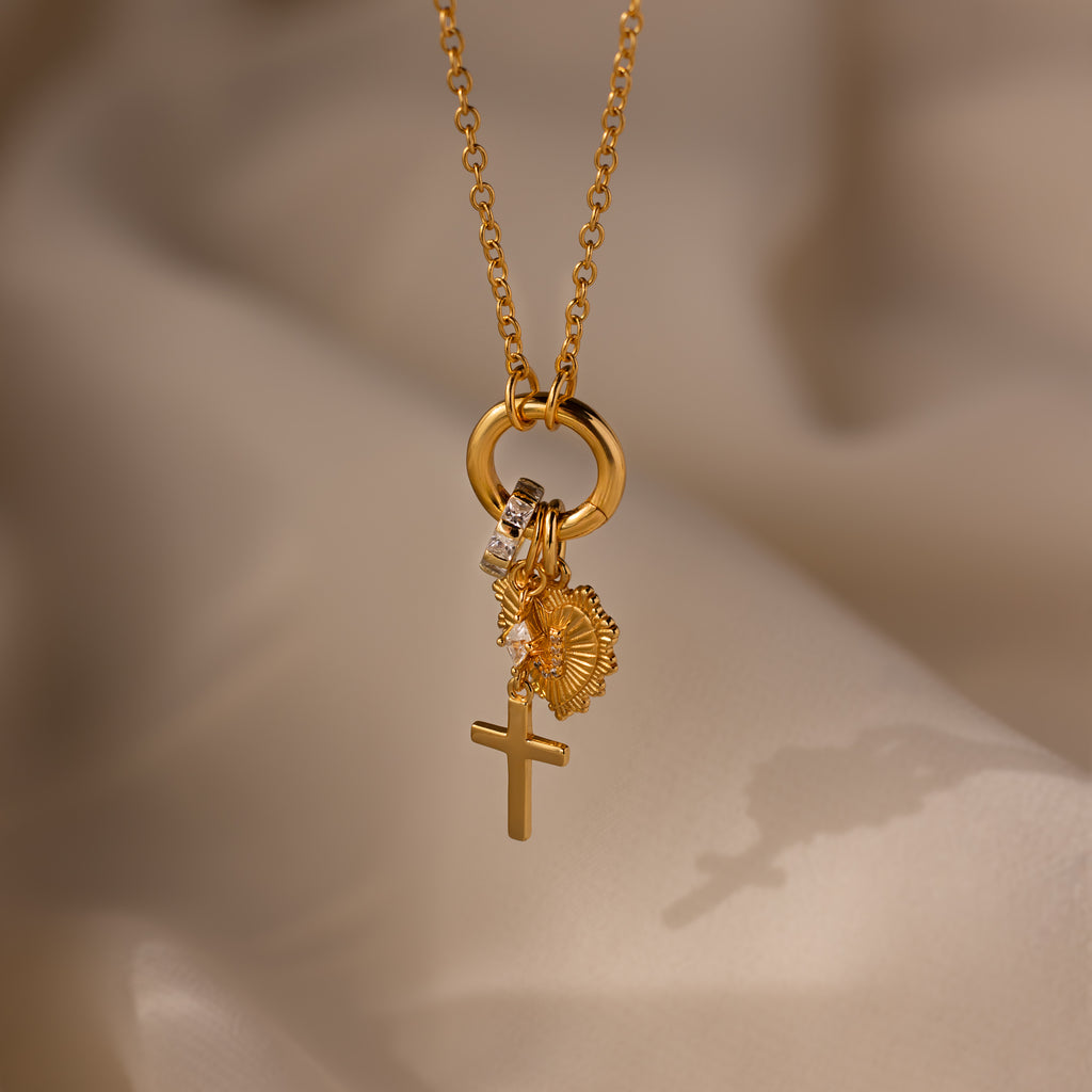 The Diamond Drop Cross Charm necklace features a gold cross pendant with flower and crystal accents, highlighted by a subtle diamond detail, displayed on soft beige fabric.
