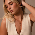A woman with long blonde hair wears a cream knit top and layered gold necklaces, including the Diamond Drop Cross Charm in 18K Gold, as she looks down thoughtfully.