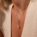 Close-up of layered gold necklaces on a woman, showcasing the Diamond Drop Cross Charm in 18K Gold with diamond accents, styled over a cream knit top.