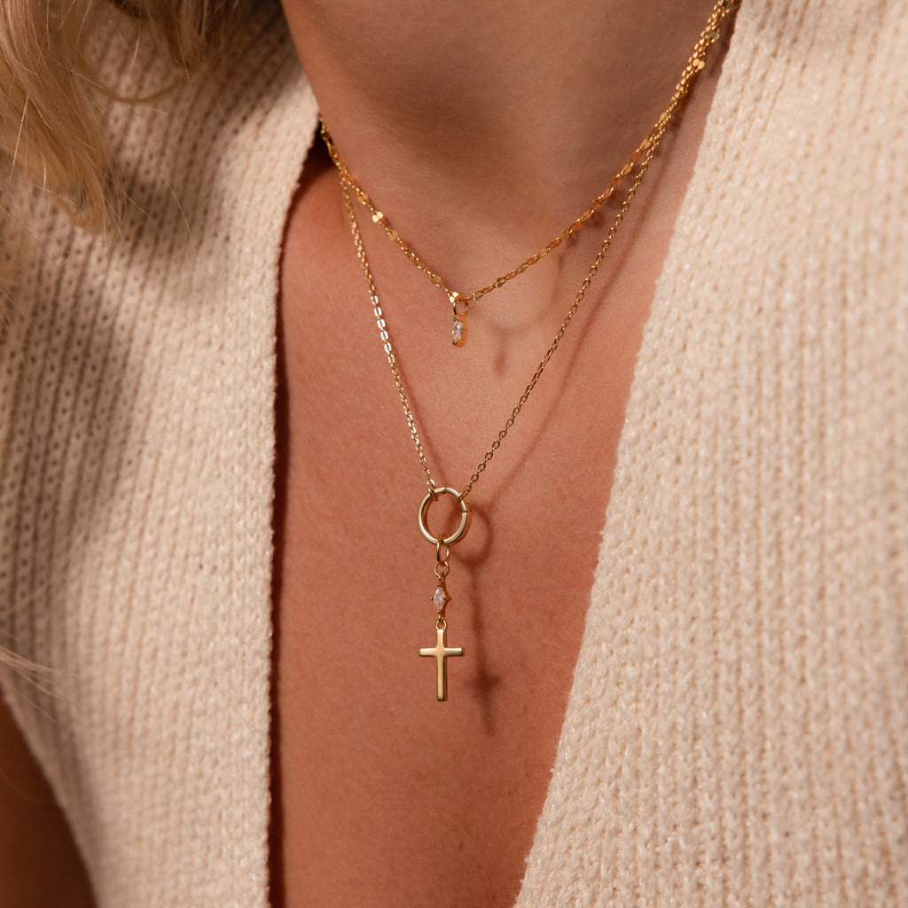A close-up shows layered gold necklaces featuring the Diamond Drop Cross Charm, styled over a cream knit V-neck top.