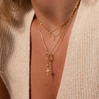 A close-up shows layered gold necklaces featuring the Diamond Drop Cross Charm, styled over a cream knit V-neck top.