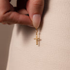 A hand displays the Diamond Drop Cross Charm in 18K Gold, featuring a subtle diamond accent, against a beige textured fabric background.