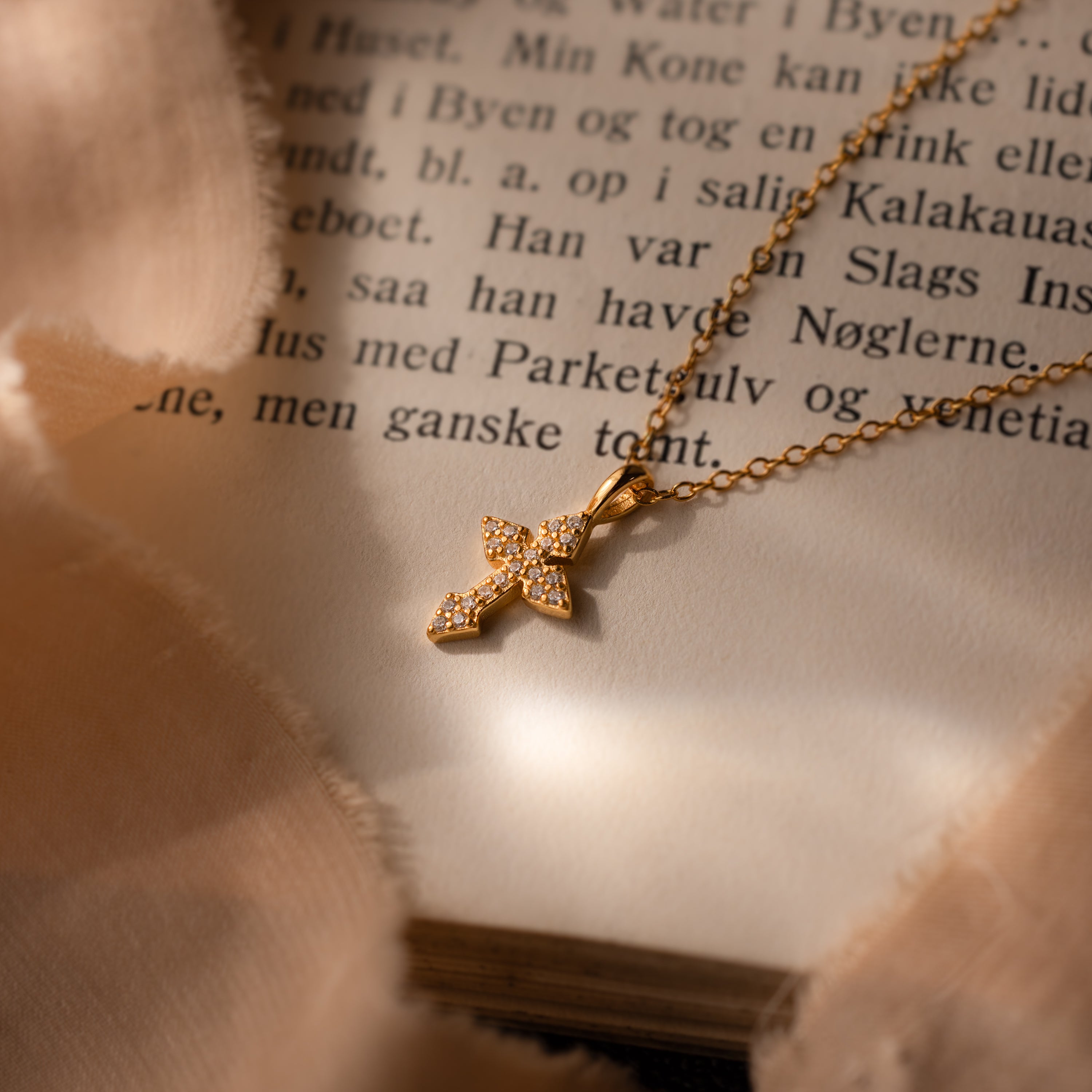 The Vintage Cross Necklace rests on an open book with Danish text, featuring a pavé finish and small diamonds.