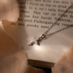 The Vintage Cross Necklace in Silver, featuring a pavé finish and delicate diamonds, rests on the open pages of a book, surrounded by soft fabric.