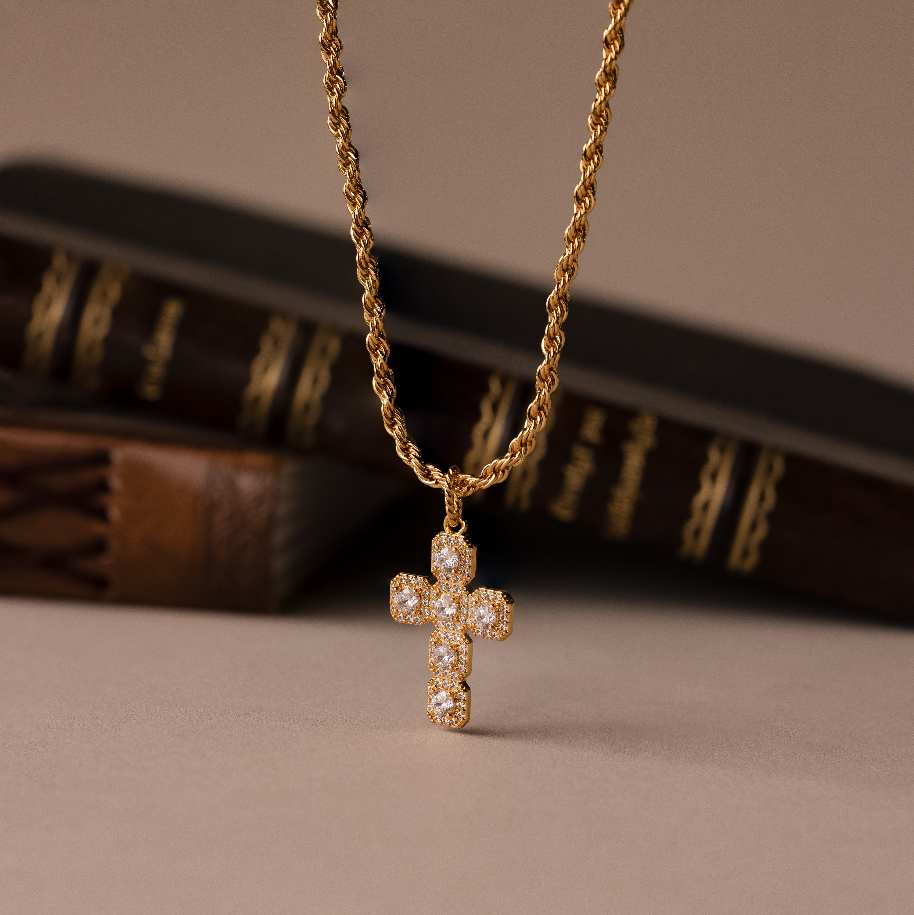 The Majesty Cross Necklace features a gold chain and diamond-studded cross pendant, showcased before vintage books—ideal for those seeking elegant faith jewelry.