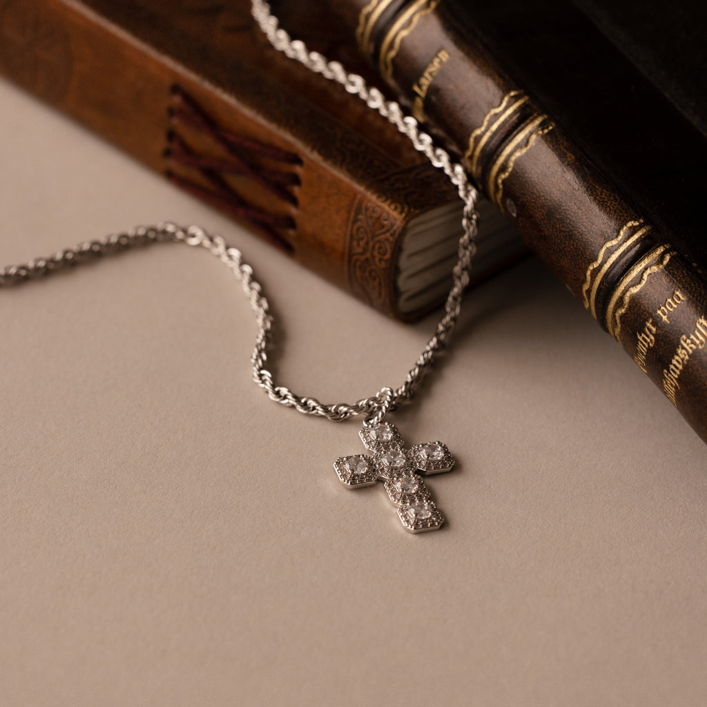 The Majesty Cross Necklace in Silver, adorned with sparkling stones, is displayed next to two vintage books on a beige surface.