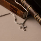 The Majesty Cross Necklace, adorned with sparkling diamonds, rests on vintage leather-bound books atop a beige surface, beautifully highlighting the enduring elegance of faith-inspired jewelry.