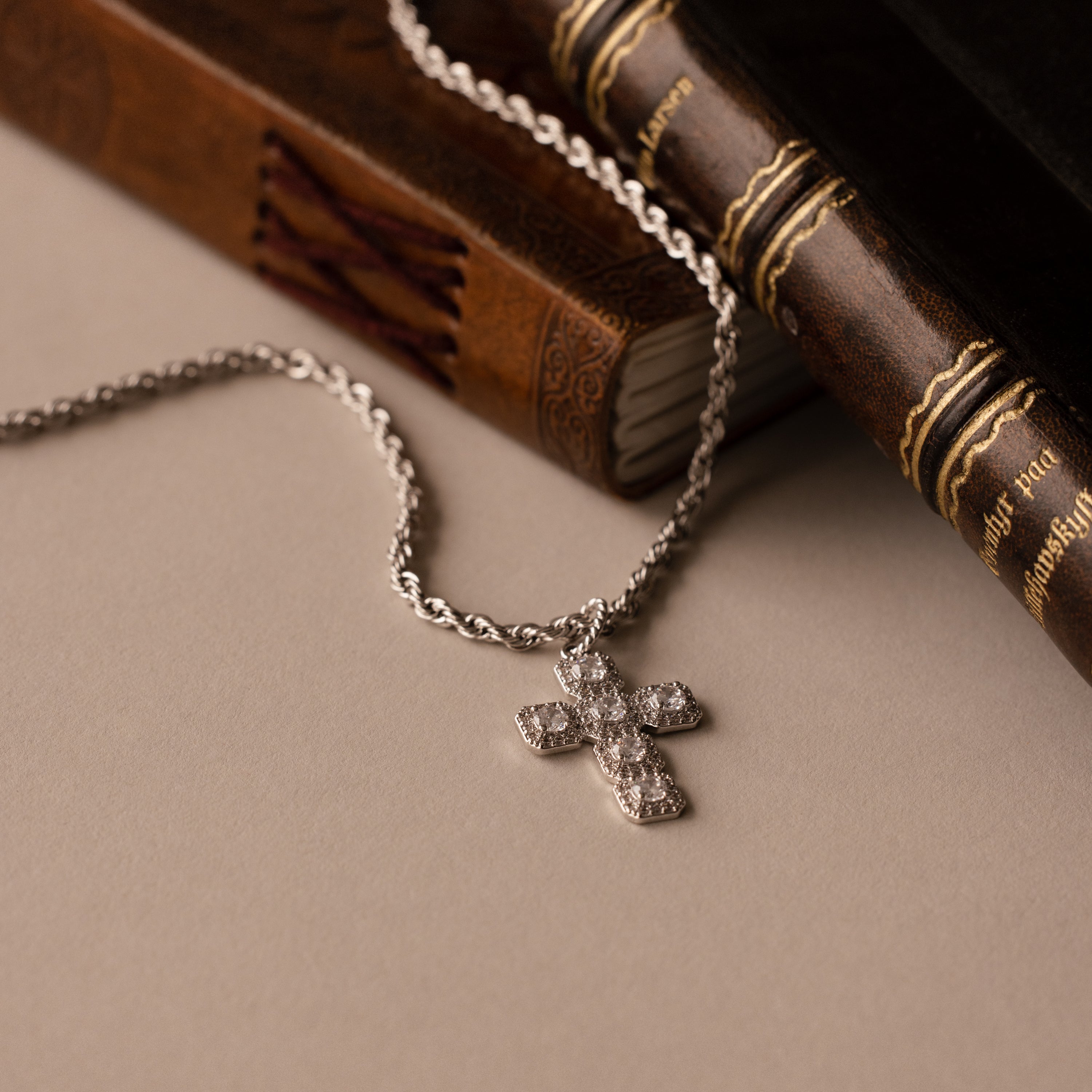 The Majesty Cross Necklace, adorned with sparkling diamonds, rests on vintage leather-bound books atop a beige surface, beautifully highlighting the enduring elegance of faith-inspired jewelry.