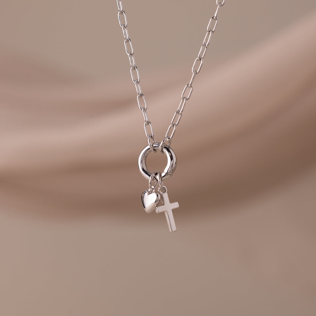 The Cross & Heart Charm Bracelet in Silver is displayed on a soft beige background.