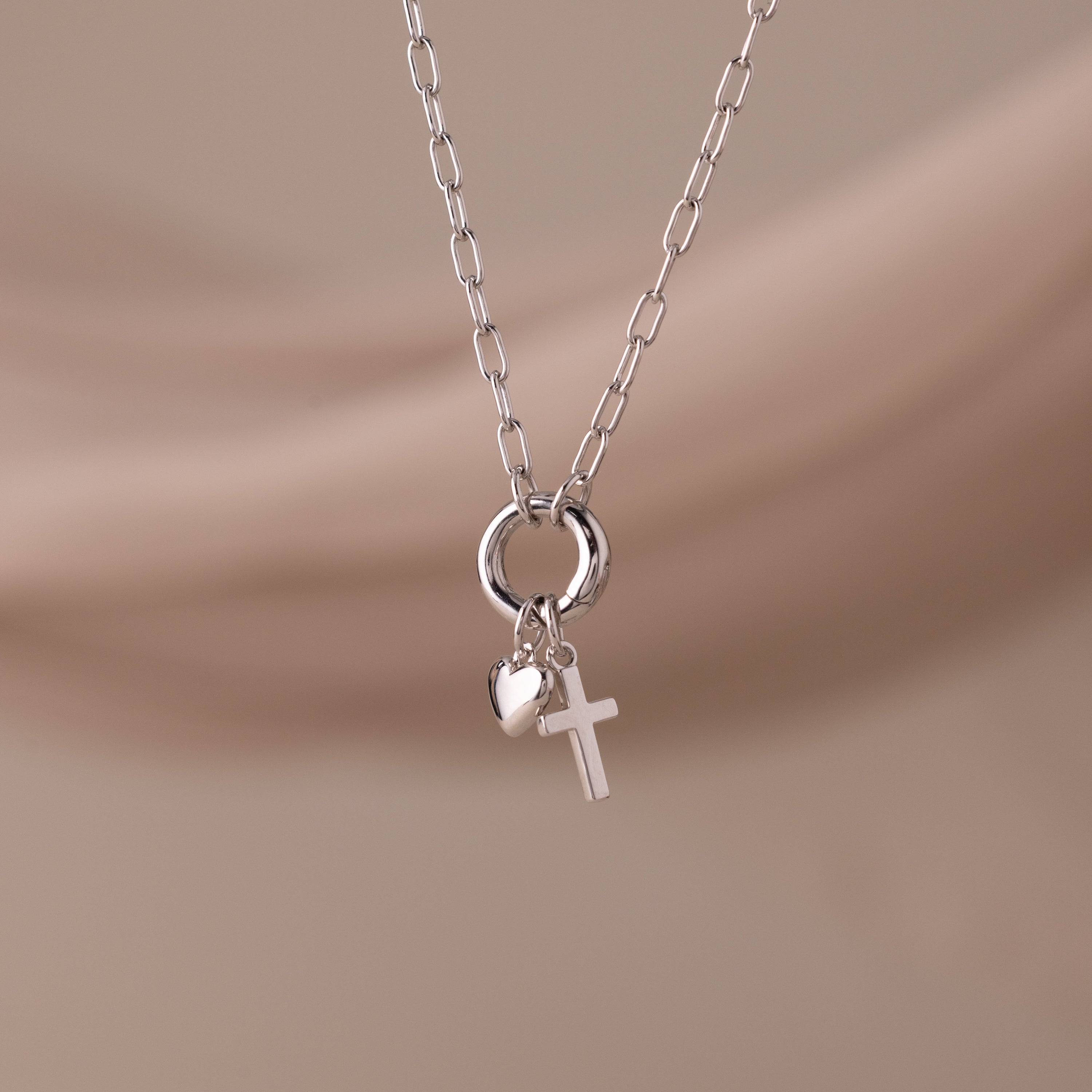 The Cross & Heart Charm Bracelet in Silver is displayed on a soft beige background.
