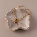 The Cross & Heart Charm Bracelet in 18K Gold is displayed in a small, speckled ceramic dish.