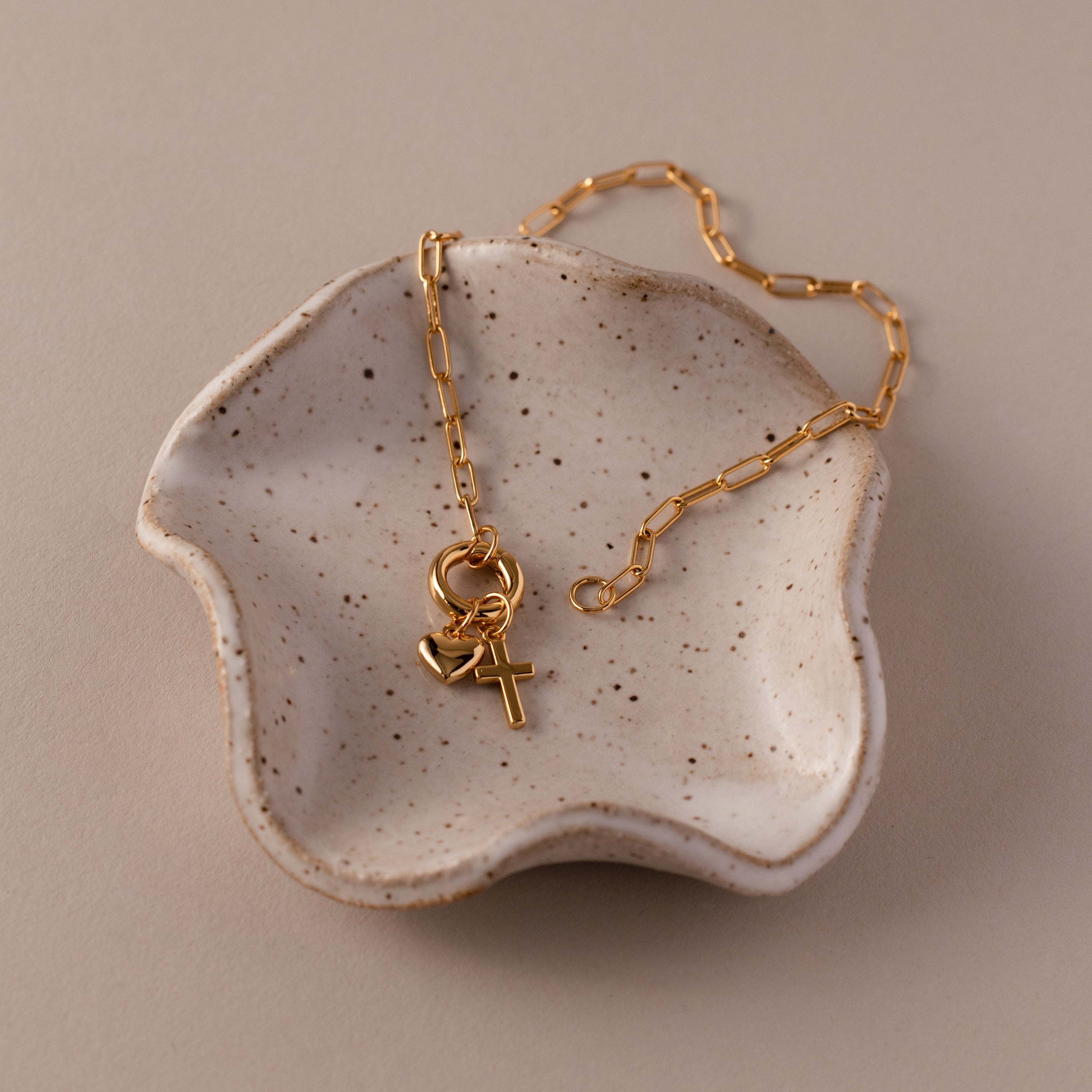 The Cross & Heart Charm Bracelet in 18K Gold is displayed in a small, speckled ceramic dish.