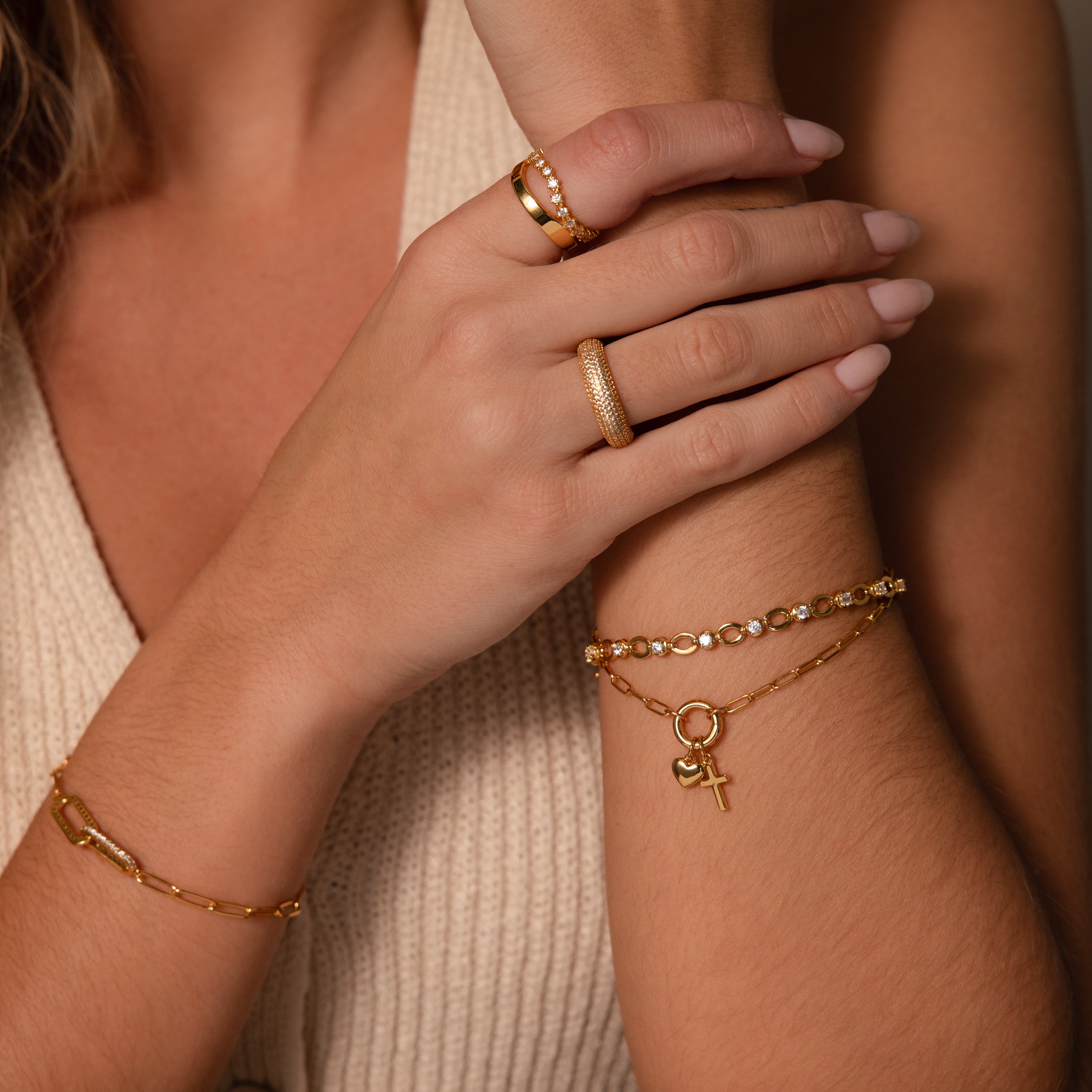 A woman’s hands showcase a Cross & Heart Charm Bracelet in 18K Gold and gold rings, styled against a beige sleeveless top.