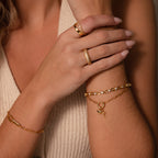 Close-up of crossed arms styled with gold rings, a minimalist bracelet, and the Cross & Heart Charm Bracelet, paired with a light-colored sleeveless top.