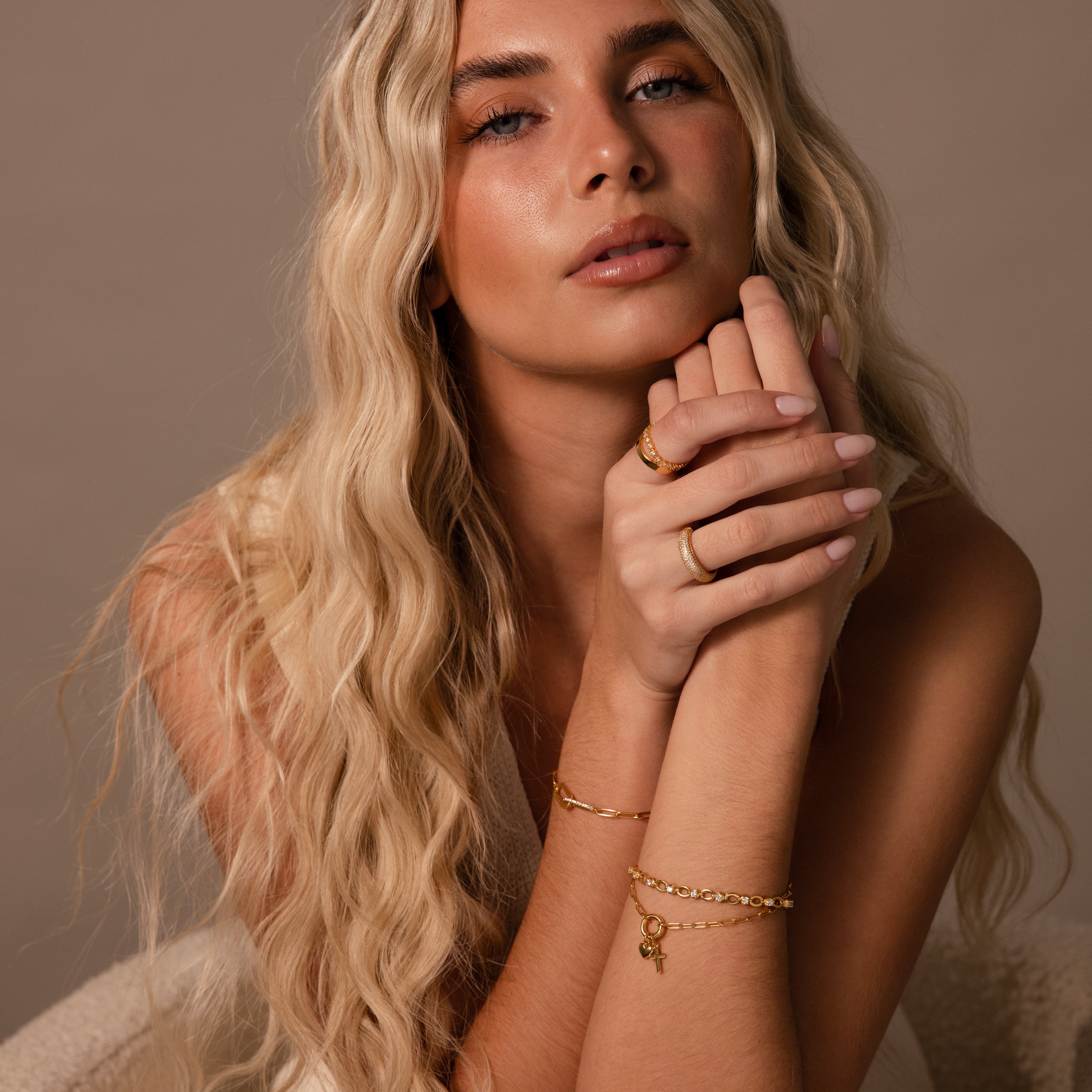 Woman with long blonde hair wearing gold jewelry, including the Cross & Heart Charm Bracelet, poses with her hands near her face against a neutral background.