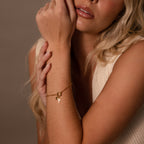A blonde woman wears the Cross & Heart Charm Bracelet in 18K Gold, its minimalist paperclip chain with cross and heart charms resting elegantly on her wrist as she touches her face thoughtfully.