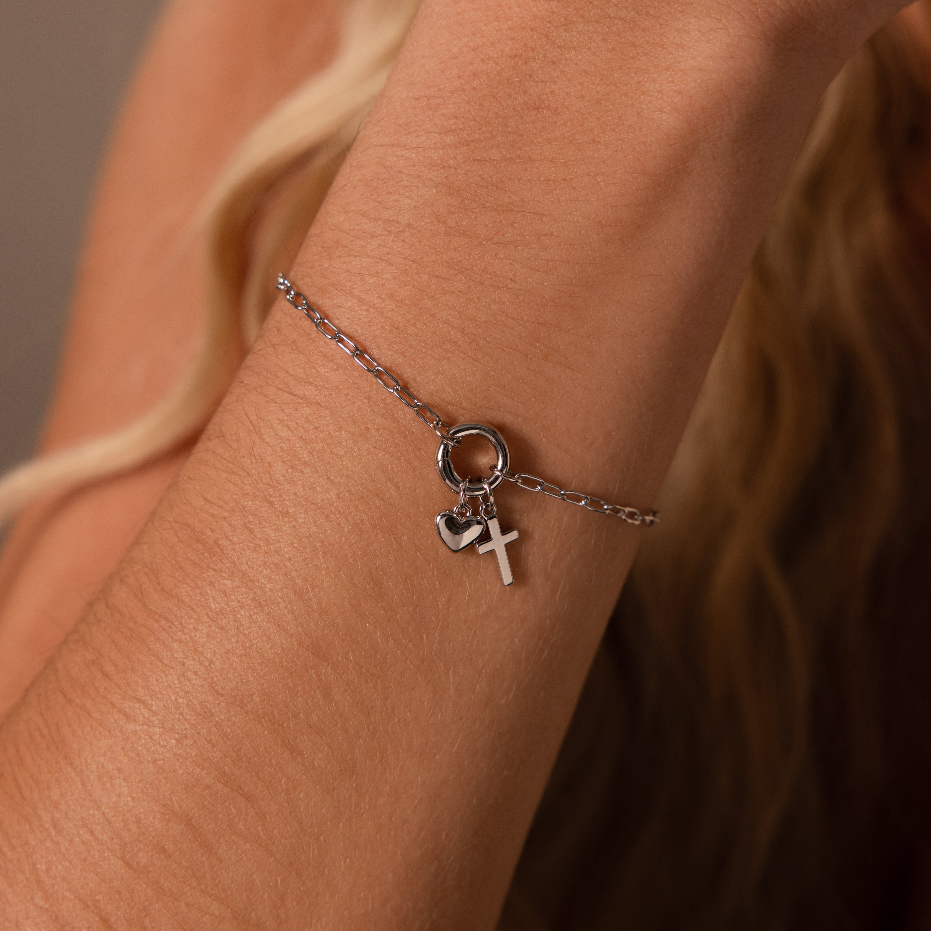 The Cross & Heart Charm Bracelet in Silver features a minimalist paperclip chain adorned with heart and cross charms, elegantly resting on the wrist.