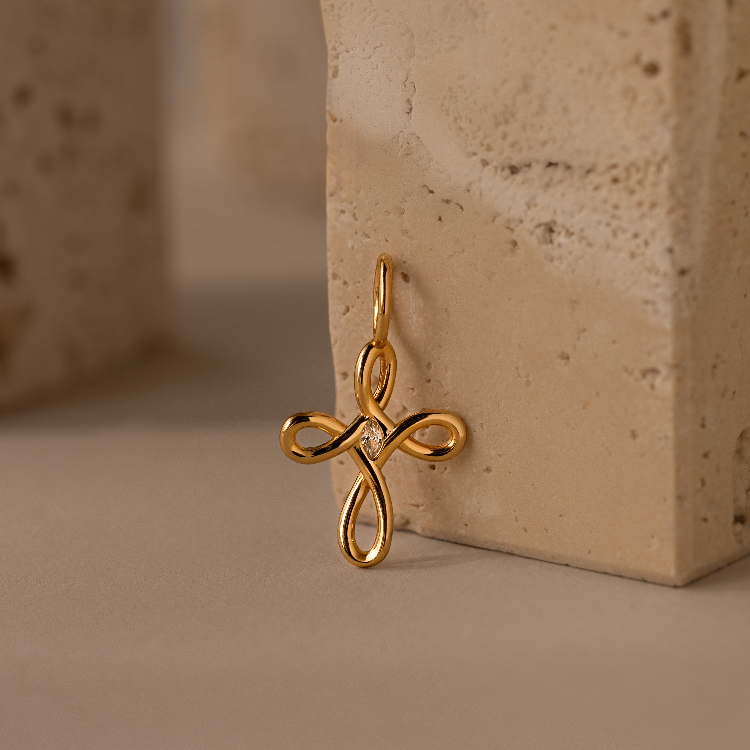 Infinity Cross Charm in 18K Gold featuring a small central gem, shown against a textured beige stone background. A meaningful piece, perfect for symbolizing eternal faith.