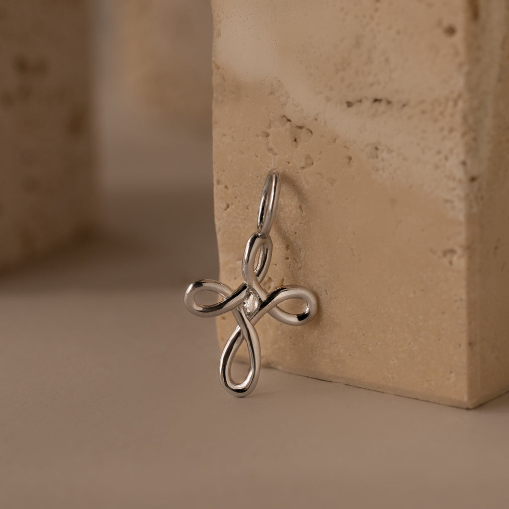 The Infinity Cross Charm in Silver, symbolizing eternal faith, rests against a beige textured stone surface.