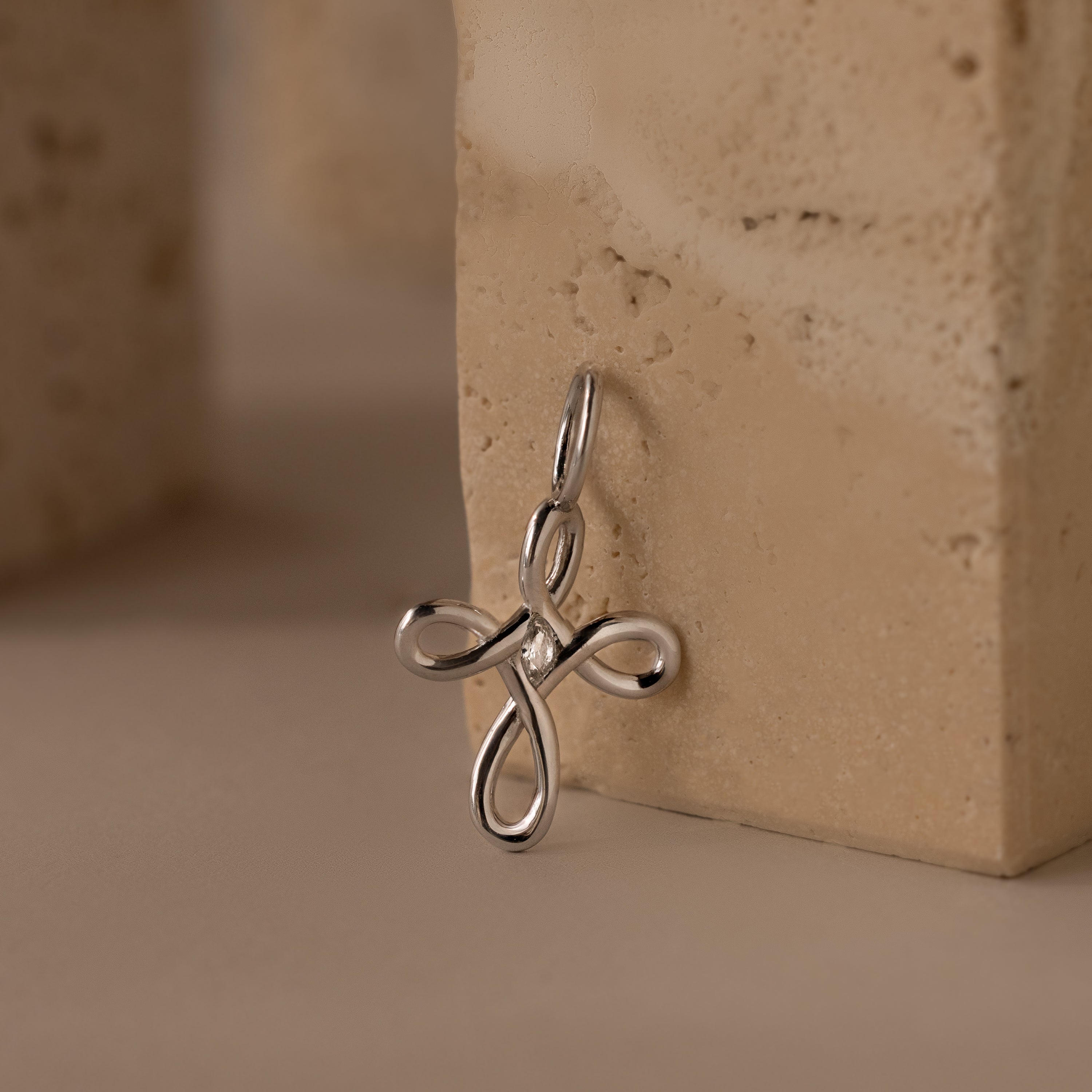 The Infinity Cross Charm in Silver, symbolizing eternal faith, rests against a beige textured stone surface.
