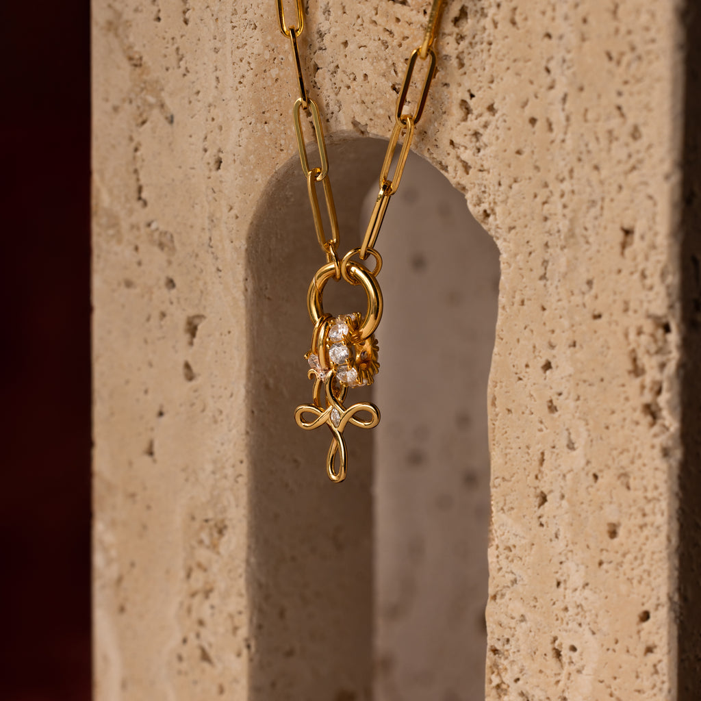The Infinity Cross Charm in 18K Gold, paired with a paperclip chain and lion charm, is elegantly displayed on a textured stone—a symbol of eternal faith and meaningful style.