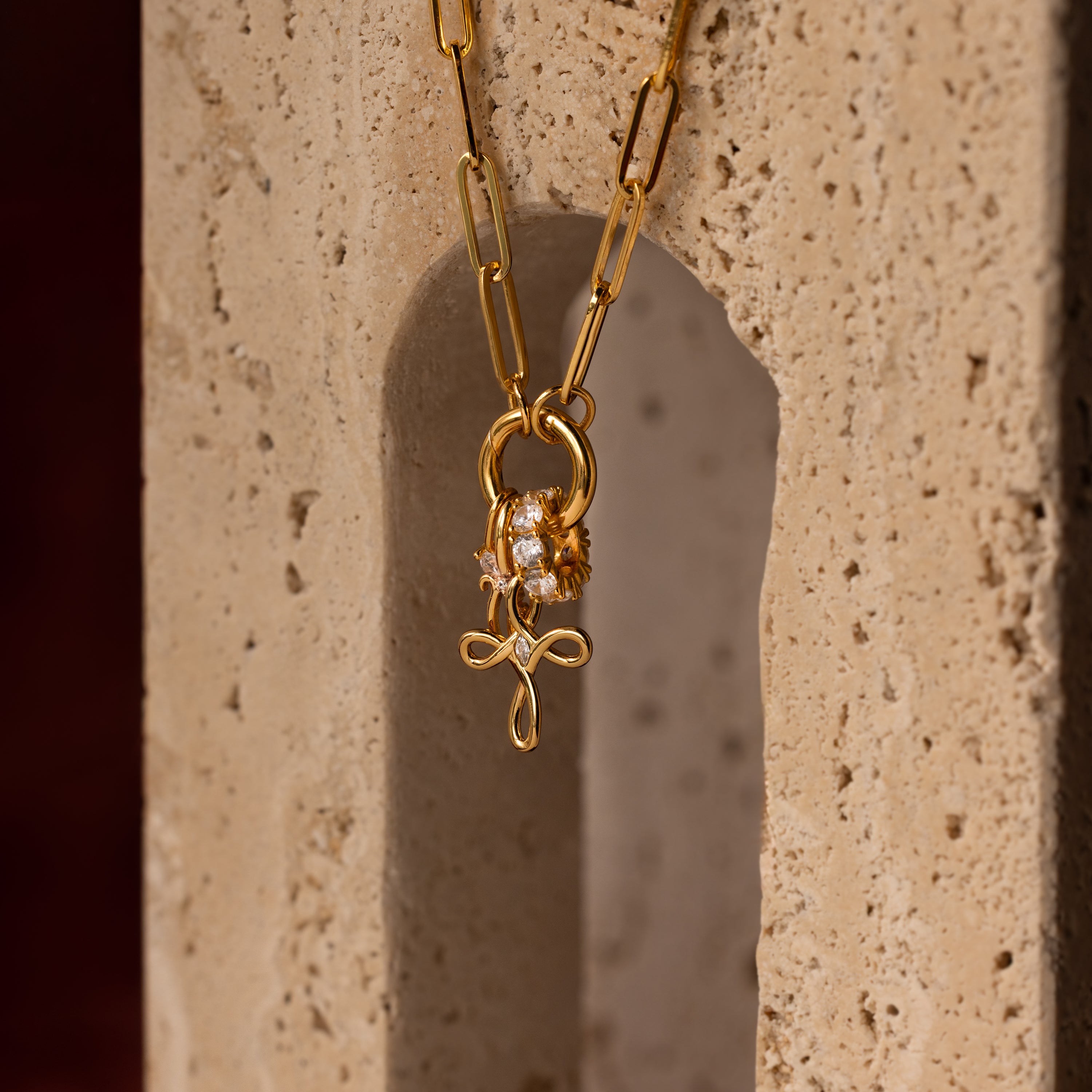 The Infinity Cross Charm in 18K Gold, paired with a paperclip chain and lion charm, is elegantly displayed on a textured stone—a symbol of eternal faith and meaningful style.