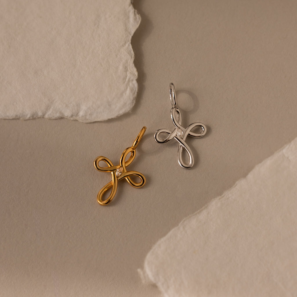 The Infinity Cross Charm, a gold and silver cross-shaped pendant, rests on a textured beige paper background—a meaningful everyday accessory that celebrates eternal faith.
