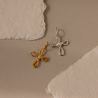 The Infinity Cross Charm, a gold and silver cross-shaped pendant, rests on a textured beige paper background—a meaningful everyday accessory that celebrates eternal faith.