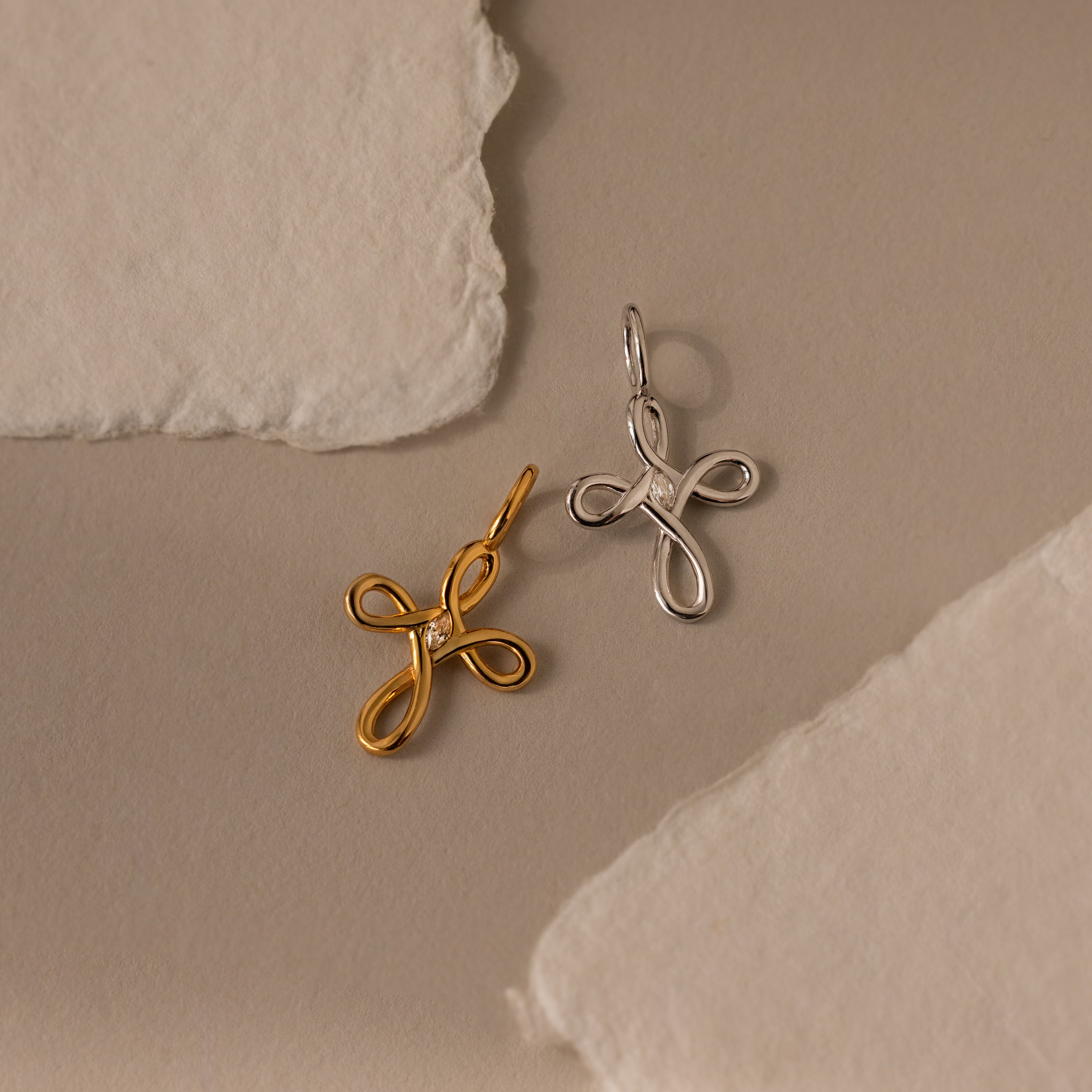 The Infinity Cross Charm, a gold and silver cross-shaped pendant, rests on a textured beige paper background—a meaningful everyday accessory that celebrates eternal faith.