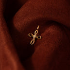 The Infinity Cross Charm in 18K Gold, featuring a looped design with a small diamond, rests on rich brown fabric—an enduring symbol of faith and timeless elegance for those who cherish meaningful jewelry.
