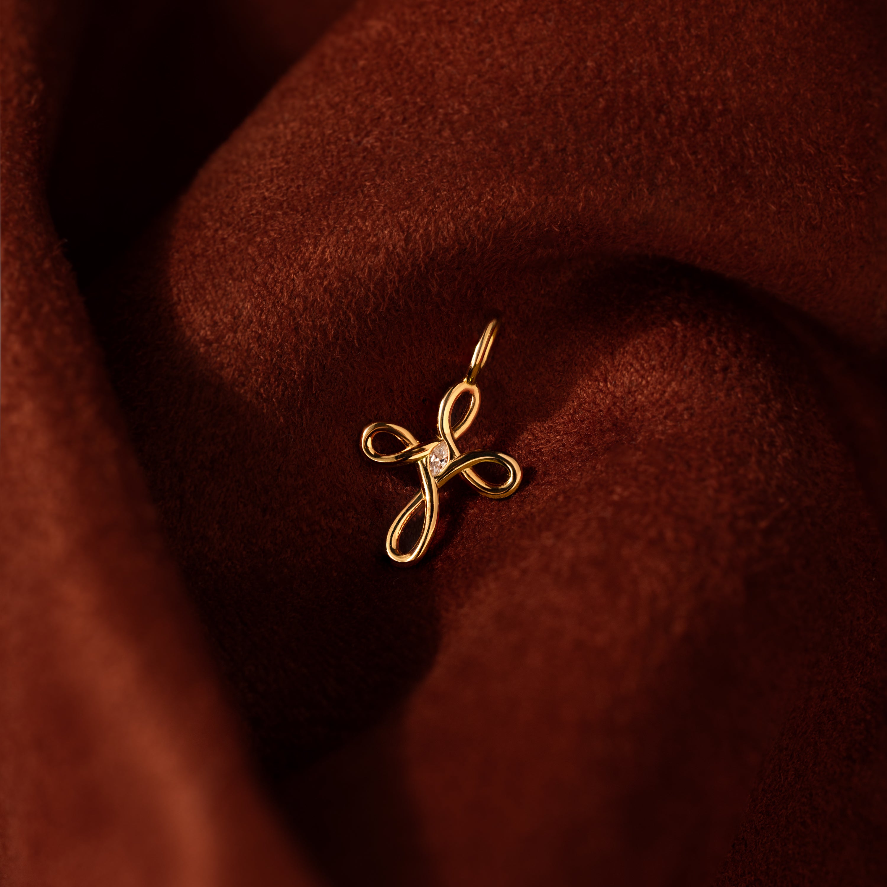 The Infinity Cross Charm in 18K Gold, featuring a looped design with a small diamond, rests on rich brown fabric—an enduring symbol of faith and timeless elegance for those who cherish meaningful jewelry.