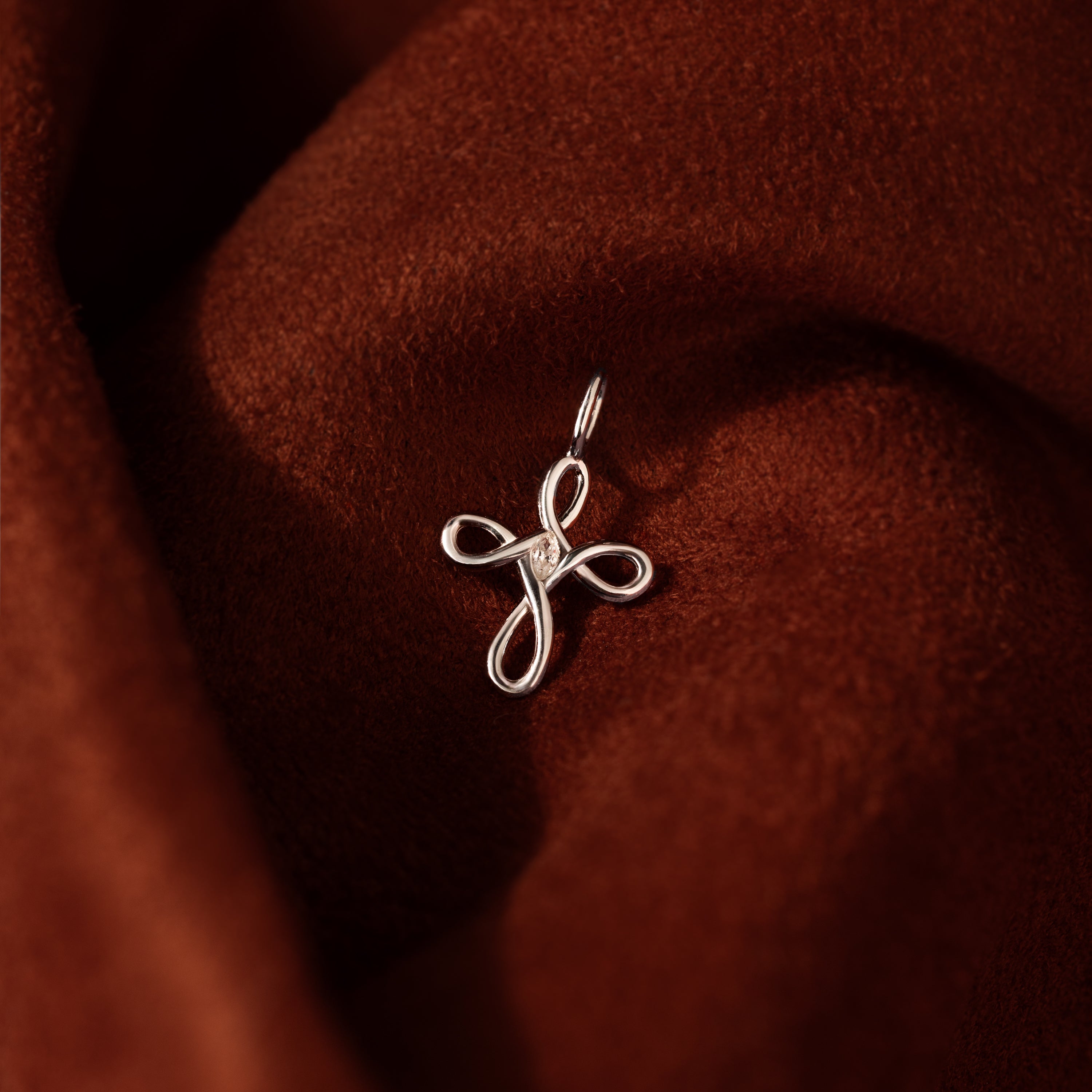 The Infinity Cross Charm, crafted in silver and symbolizing eternal faith, rests on textured brown fabric—a meaningful charm for daily wear.