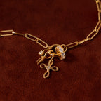 The Infinity Cross Charm in 18K Gold is shown on a brown surface, featuring exquisite jeweled accents, a floral pendant, and meaningful gold chain necklace charms.