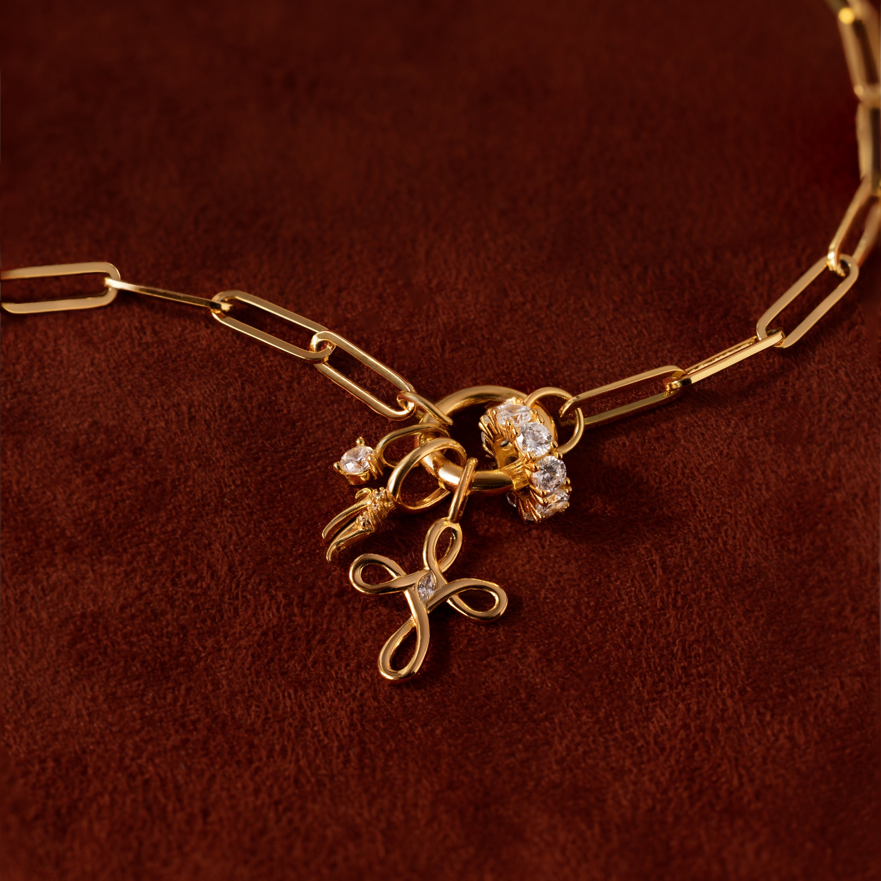 The Infinity Cross Charm necklace, featuring a gold chain and diamond floral charms, symbolizes eternal faith and is displayed on a brown velvet background.
