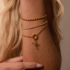 A close-up of a woman's wrist adorned with three gold bracelets, including the Infinity Cross Charm in 18K Gold symbolizing eternal faith.