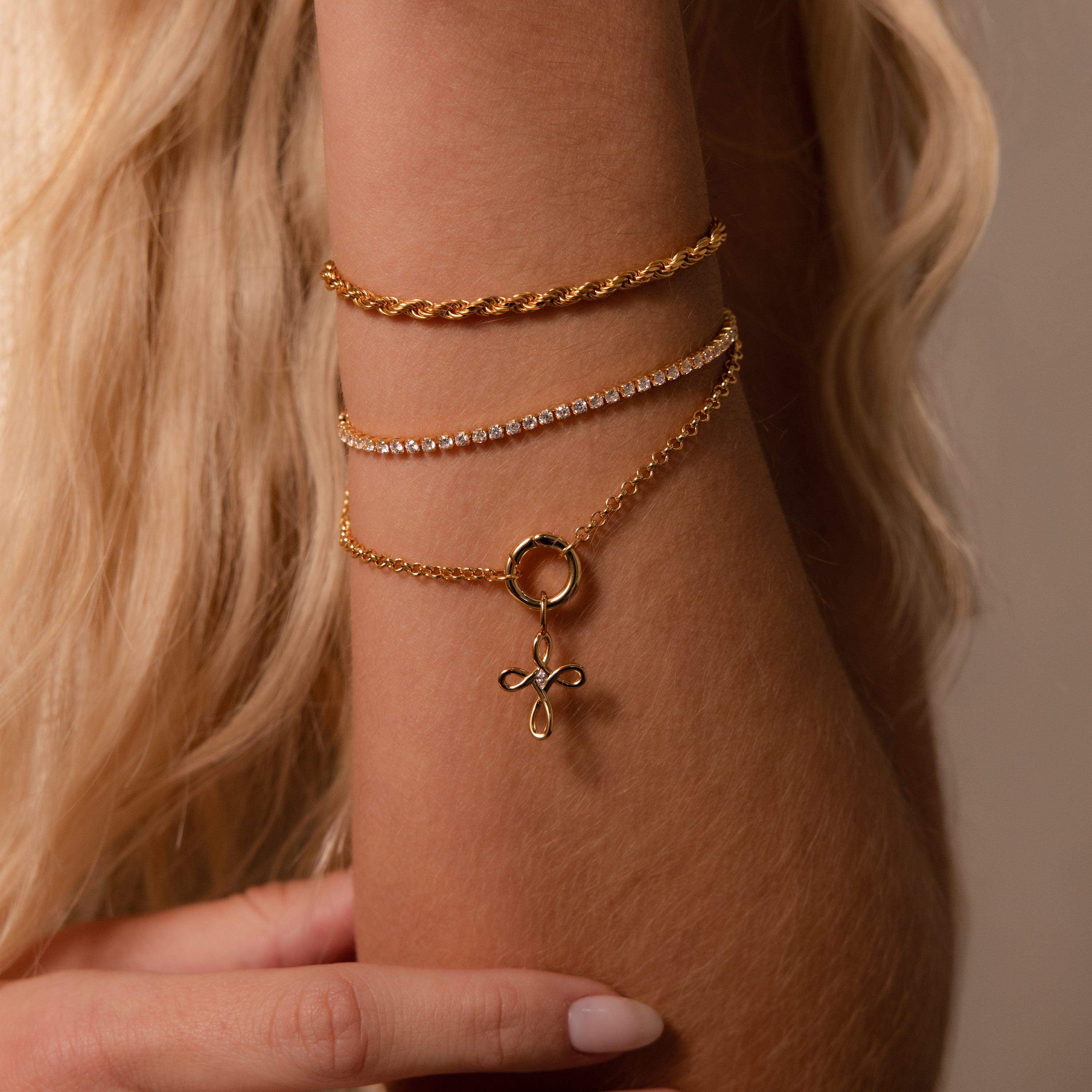 A close-up of a woman's wrist adorned with three gold bracelets, including the Infinity Cross Charm in 18K Gold symbolizing eternal faith.