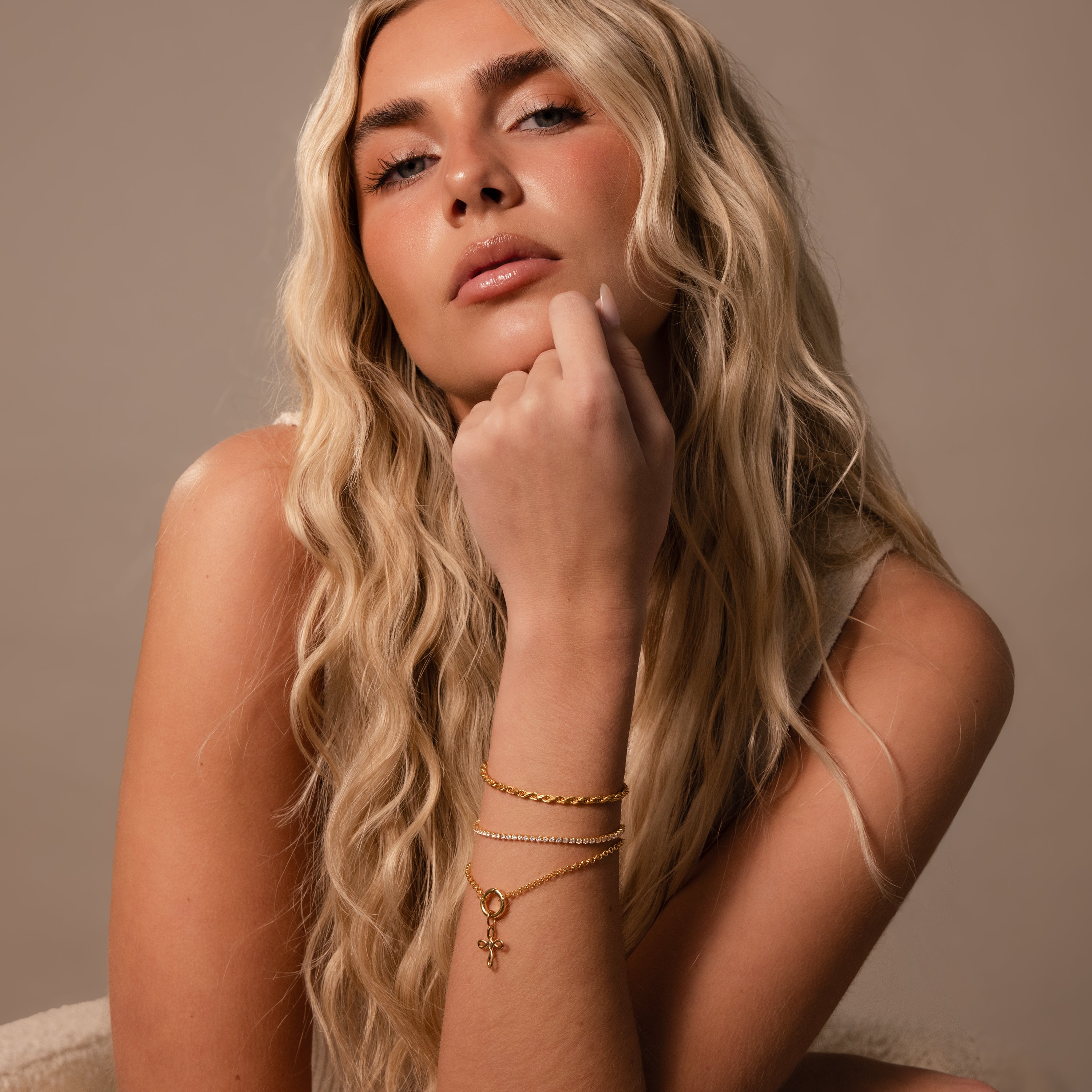 A woman with long wavy blonde hair and gold bracelets poses confidently in a sleeveless top, her look elevated by the Infinity Cross Charm in 18K Gold, adding a touch of meaningful elegance.
