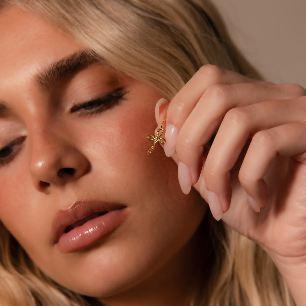 A woman with blonde hair gently holds the Infinity Cross Charm in 18K Gold near her face, eyes closed, feeling the powerful symbol of eternal faith.