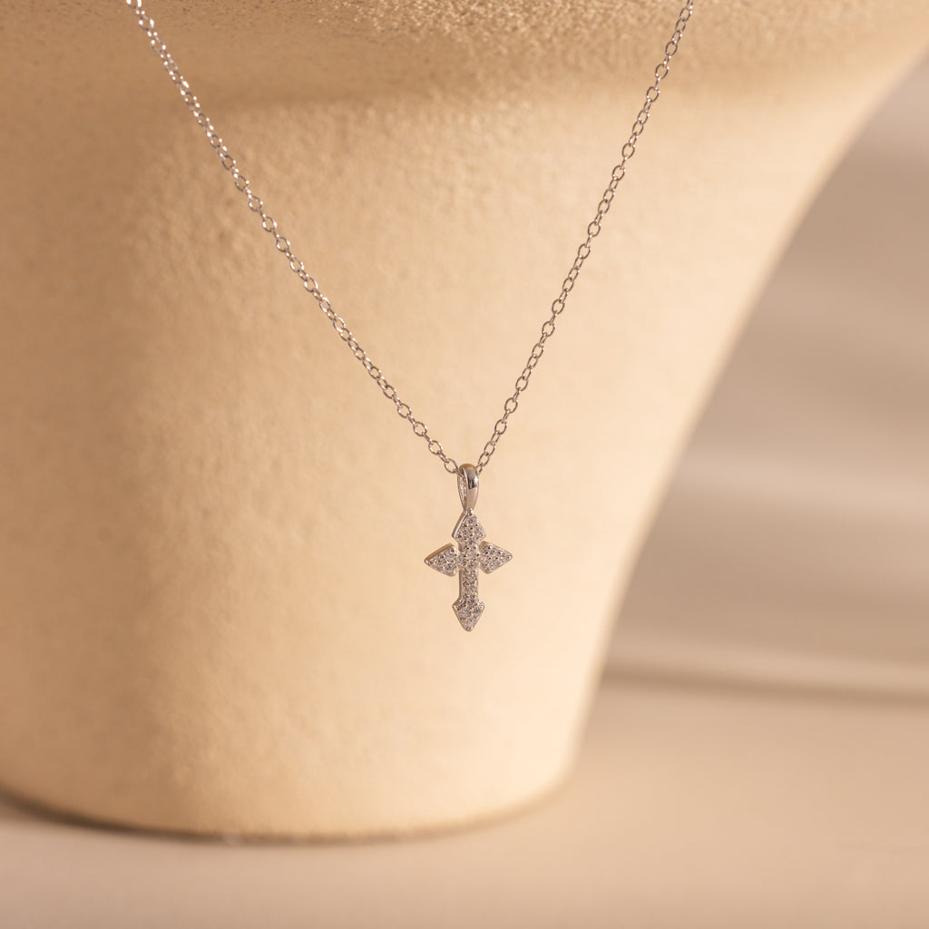 The Vintage Cross Necklace in Silver features a diamond-studded pavé cross pendant on a silver chain, shown against a beige textured background.