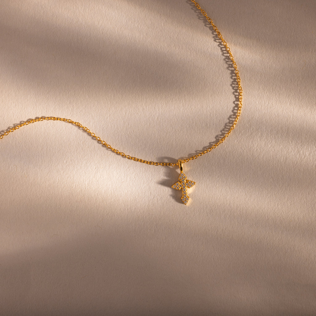 The Vintage Cross Necklace in 18K Gold features a minimalist gold chain with a small, textured pavé cross pendant, shown on a soft beige background.