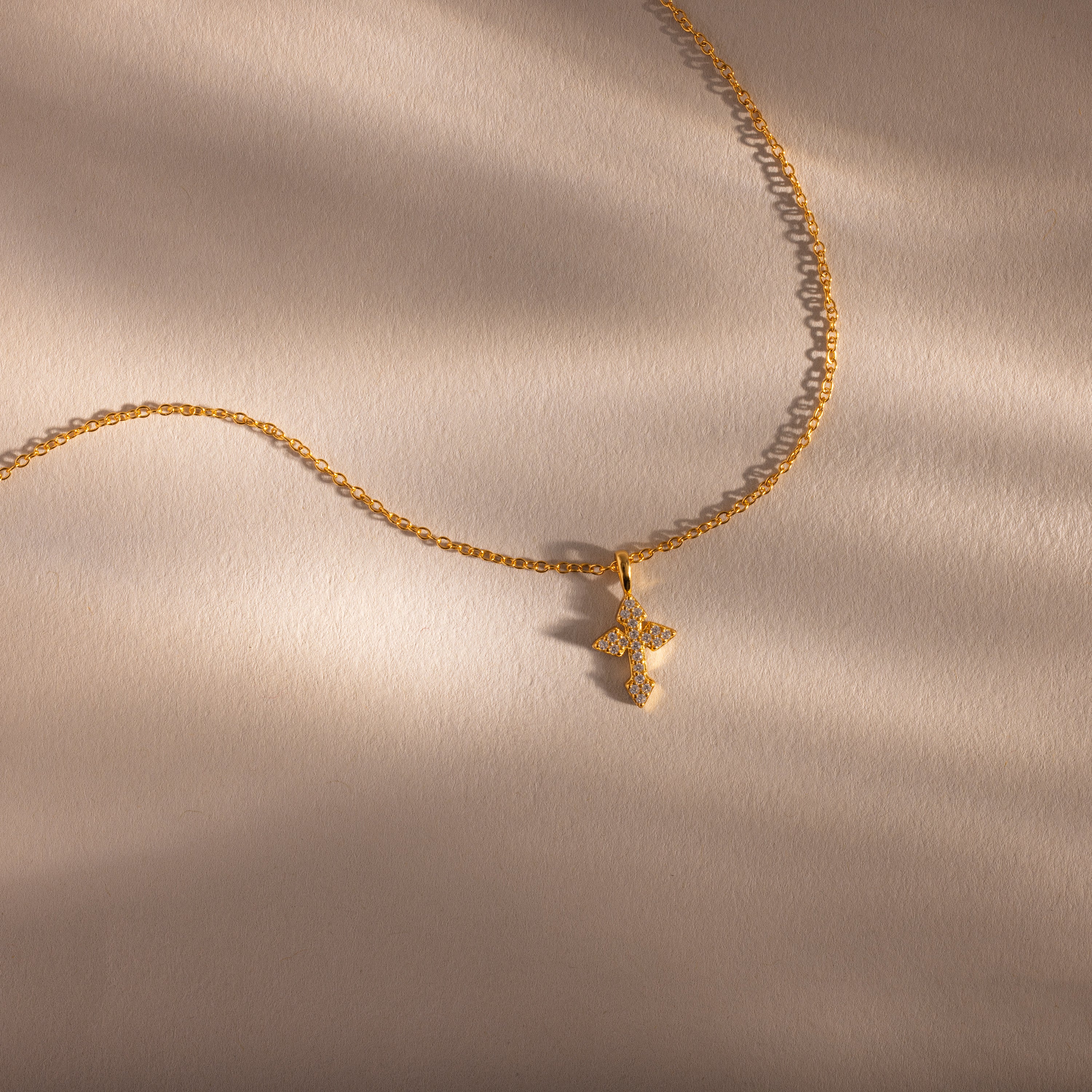 The Vintage Cross Necklace in 18K Gold features a minimalist gold chain with a small, textured pavé cross pendant, shown on a soft beige background.