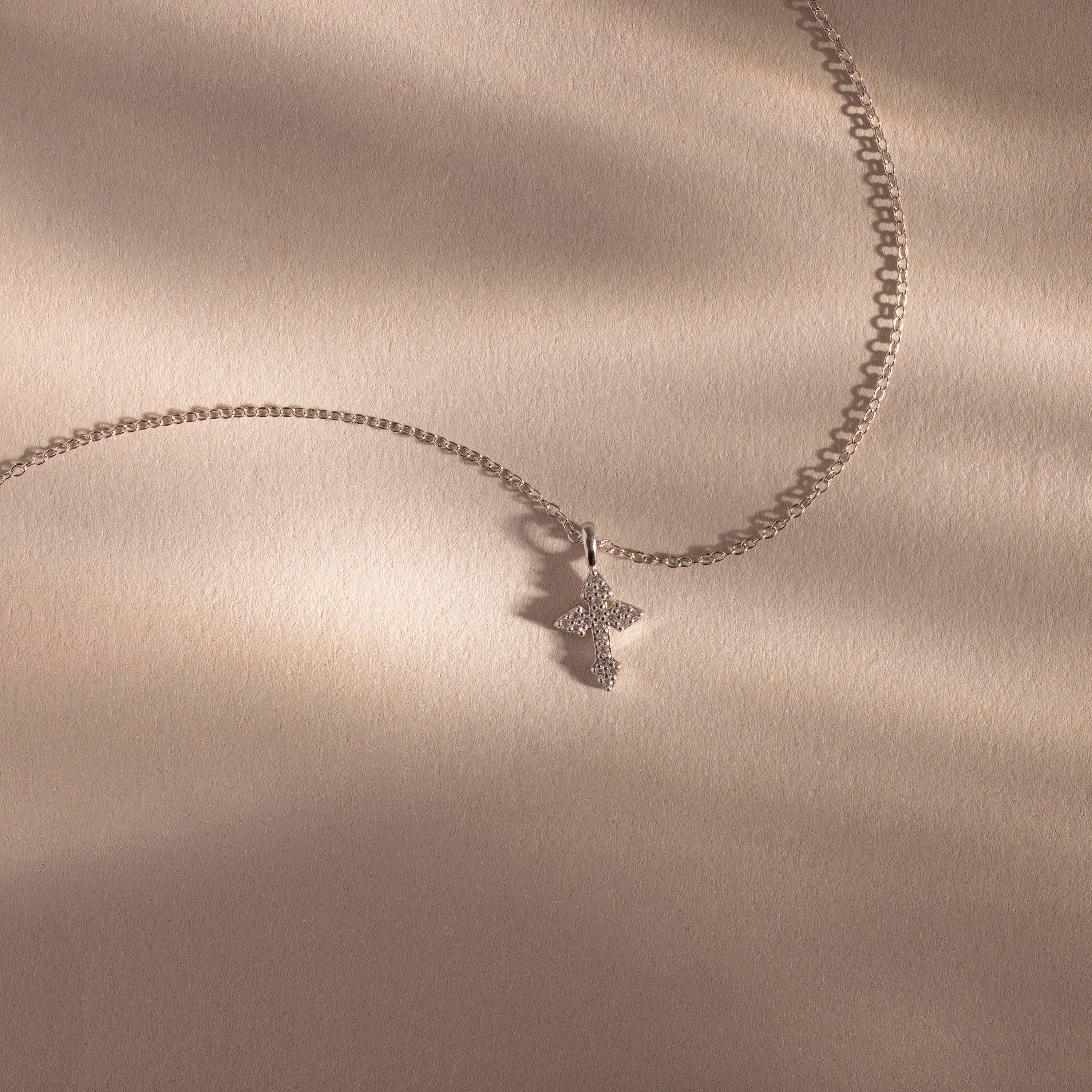 The Vintage Cross Necklace in Silver is a minimalist piece featuring a pavé-finish, diamond-studded cross pendant, shown on a beige textured surface.